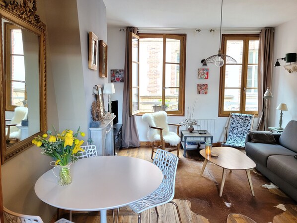 A cozy living room with a round table, a sofa, and a chair. There are two windows with curtains, a fireplace, and a mirror on the wall.