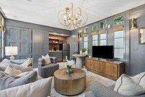 A modern living room with a grey sofa set, a round coffee table with a plant, a TV, and a wooden cabinet. - Serene Cottage with Beautiful Garden & Heated Pool (Charleston)