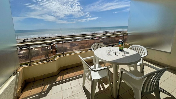 Een balkon met uitzicht op het strand en de oceaan, met een tafel, stoelen en een pak sap.