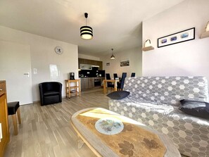 A living room with a sofa, a coffee table, a dining area with chairs, a kitchen in the background, and a clock on the wall.
