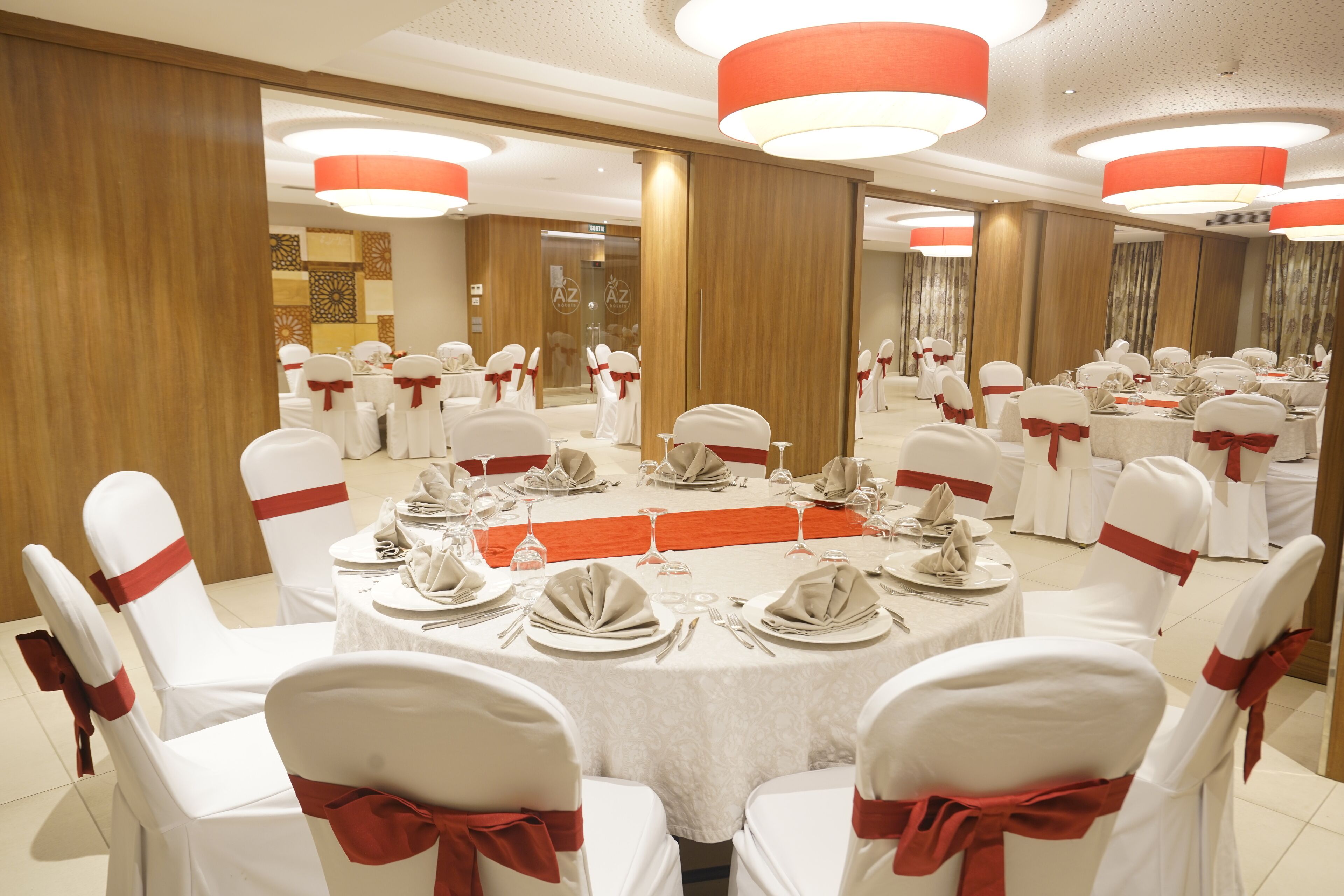 Un salón de banquetes con mesas redondas dispuestas para un evento formal, sillas blancas con cintas rojas e iluminación circular suspendida del techo.