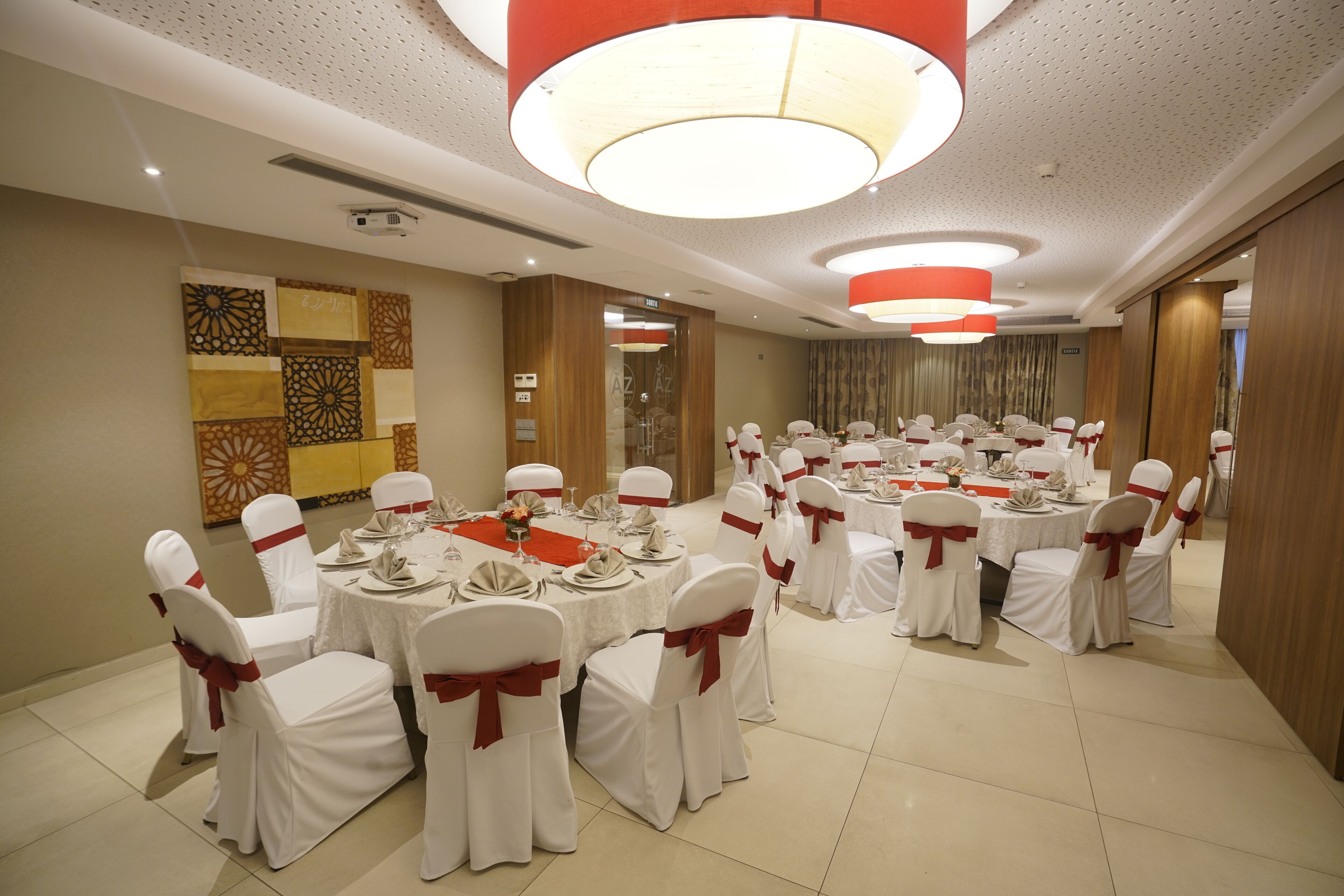 A banquet hall with round tables set for a formal event, white chairs with red sashes, and modern lighting fixtures.