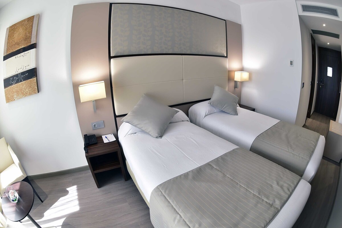 Habitación de hotel con dos camas, una mesita de noche con lámpara, una pequeña mesa con una botella y una obra de arte colgada en la pared.
