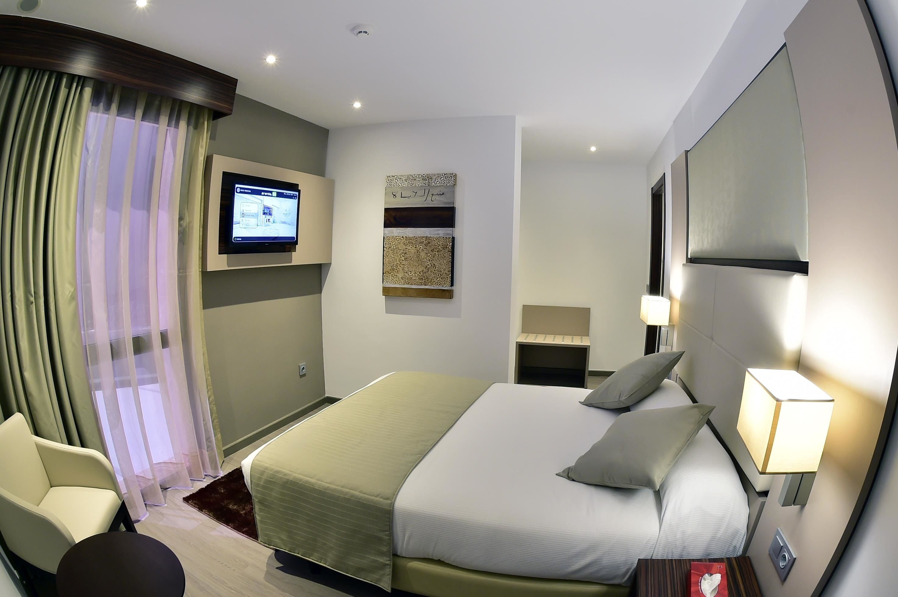 Una habitación de hotel moderna con una cama grande, un televisor montado en la pared, un escritorio con lámpara y una silla.