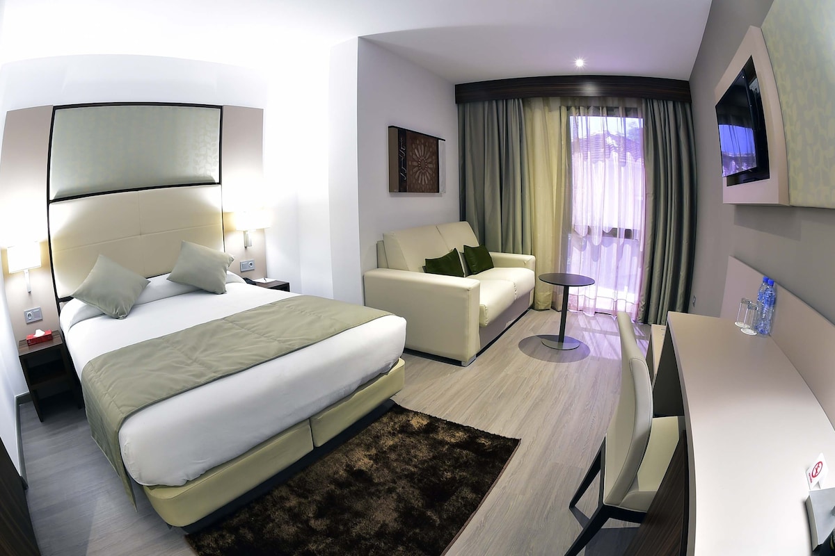 Una habitación de hotel moderna con una cama grande, un sofá, una mesita y un televisor montado en la pared.