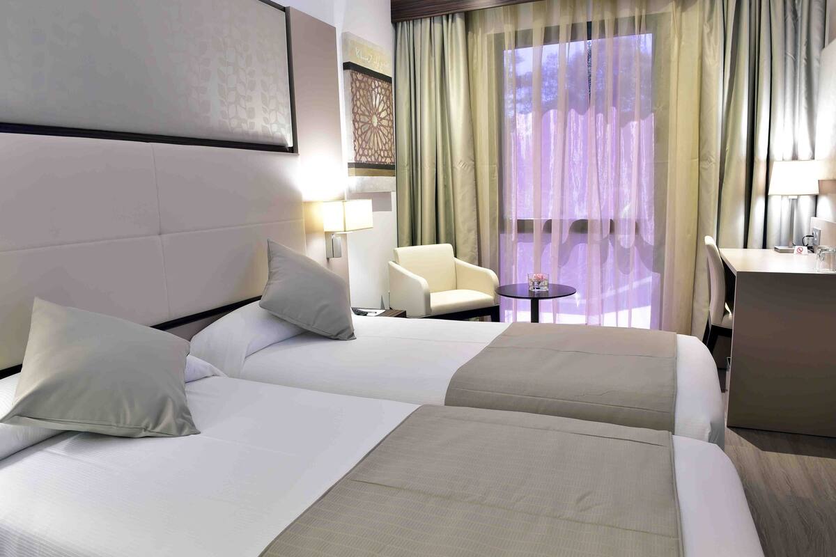 Habitación de hotel con dos camas, una silla, una mesita redonda y un escritorio con lámpara.