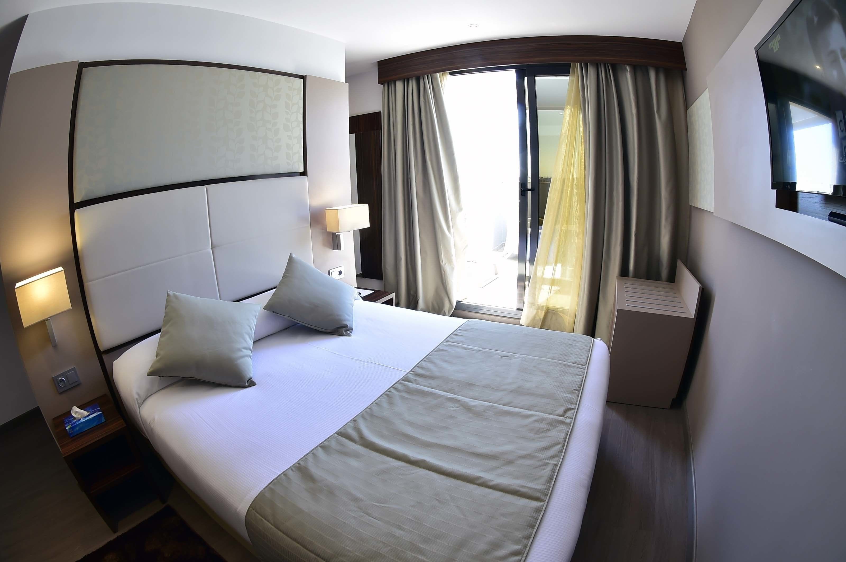 A hotel room with a large bed, a headboard, a TV mounted on the wall, a bedside table with a lamp, and a window with curtains.