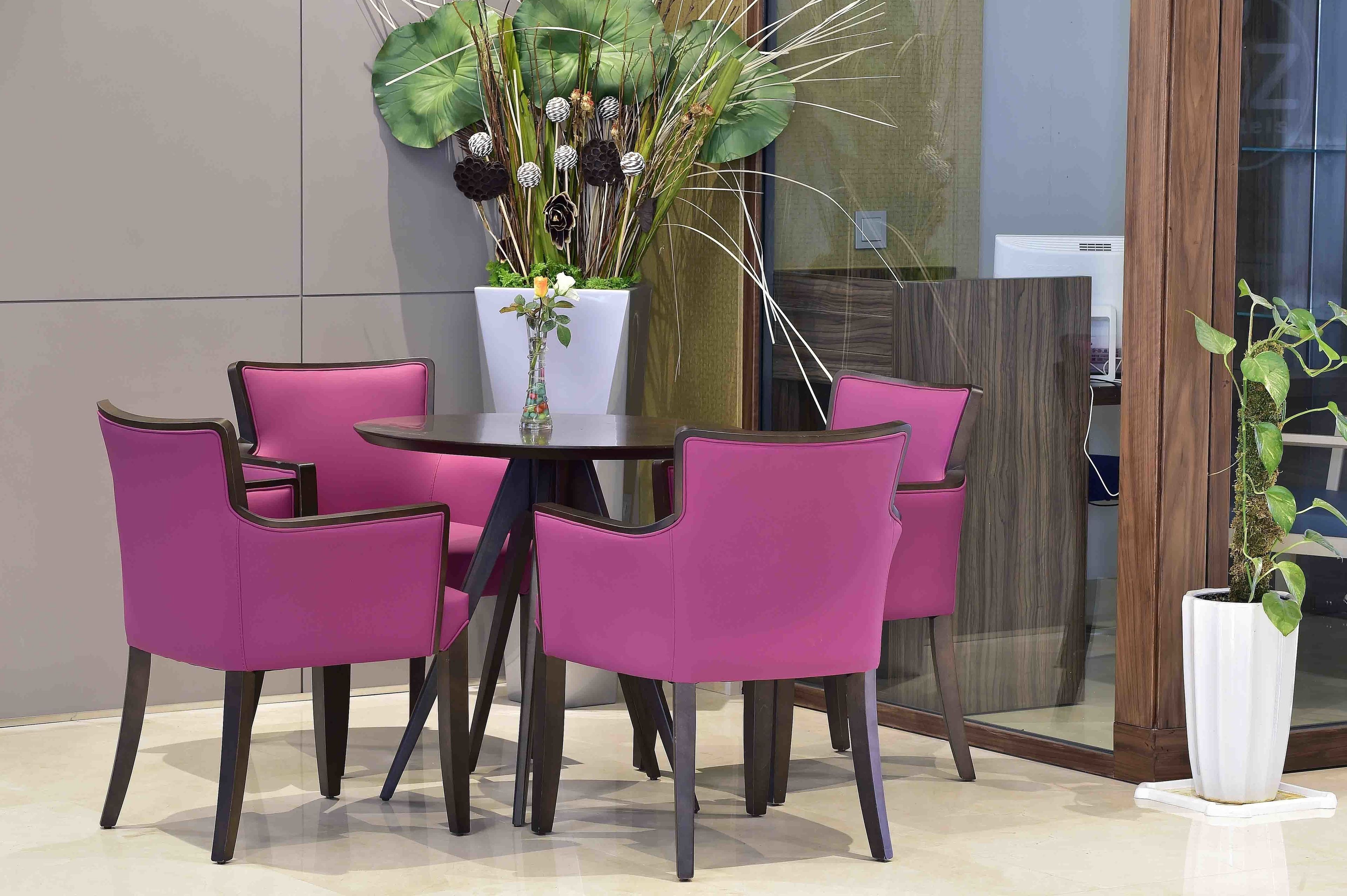 Een moderne binnensetting met een ronde tafel, omringd door vier paarse stoelen, een witte vaas met bloemen en een grote potplant met brede bladeren.