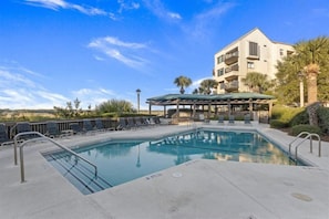 A swimming pool with lounge chairs and a covered seating area, surrounded by a building with balconies.