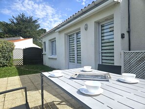A patio with a table set for two, a white building with shutters, and a fence in the background.