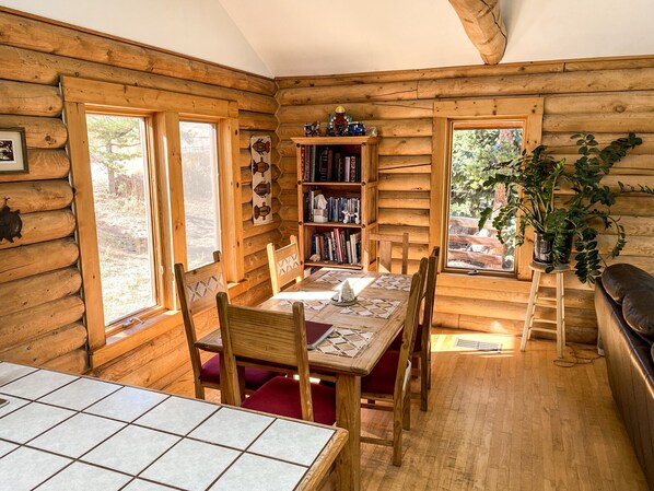 A log cabin interior with a dining table, chairs, a bookshelf, and a window.