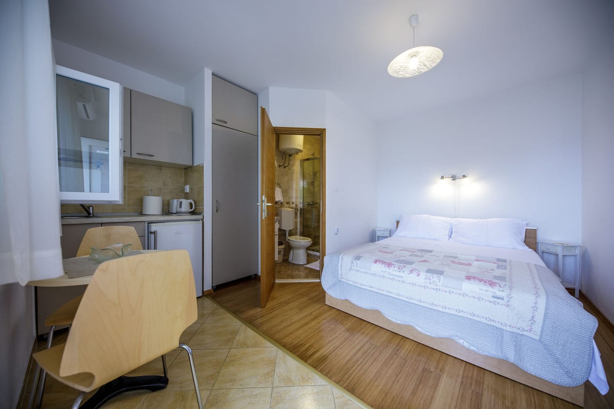 Quarto com cama, mesas de cabeceira, uma cadeira, escrivaninha e banheiro com vaso sanitário e chuveiro.