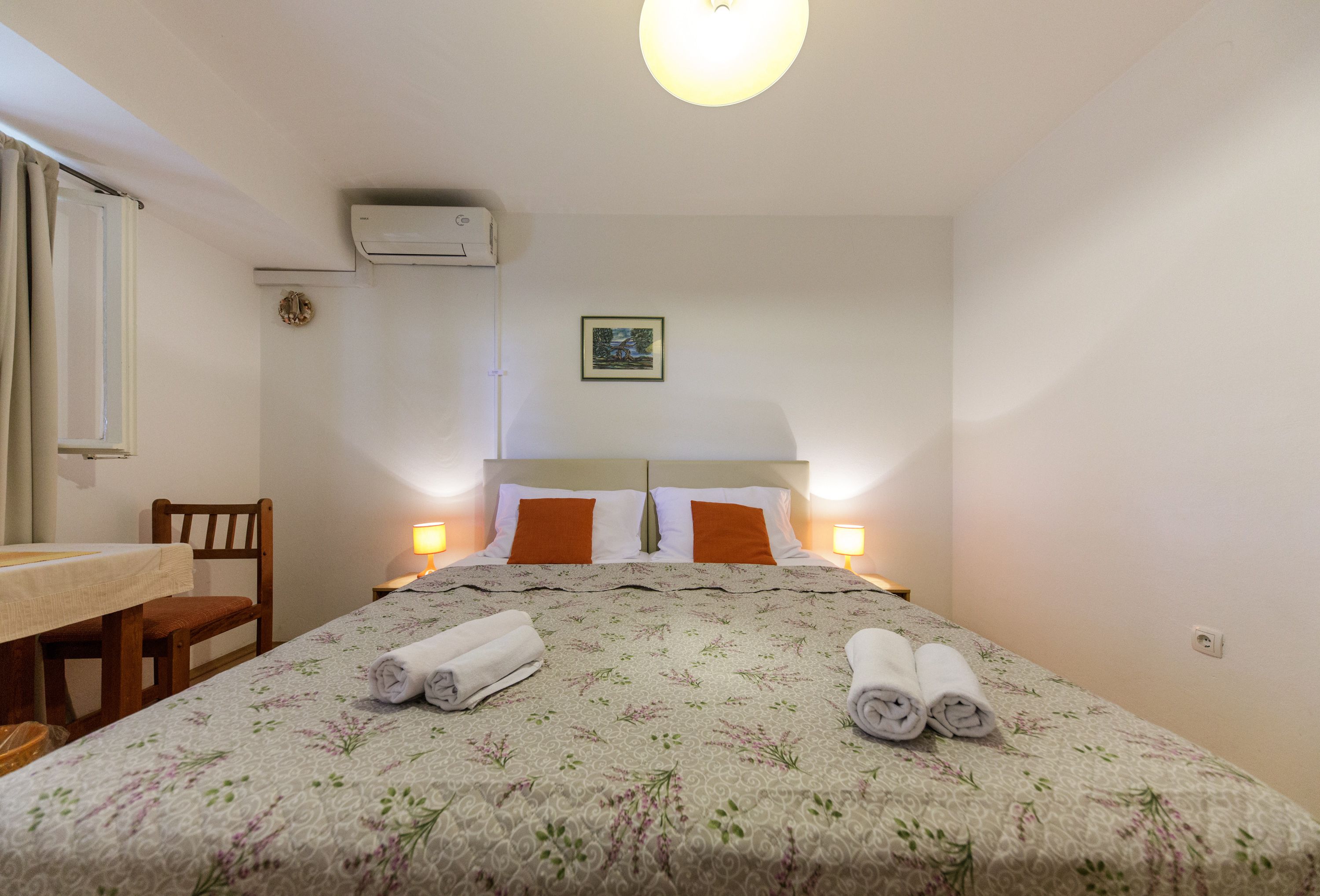 A neatly made bed with a floral bedspread, two pillows, and rolled towels on the bed. There is a wooden chair and a small table with a white tablecloth.