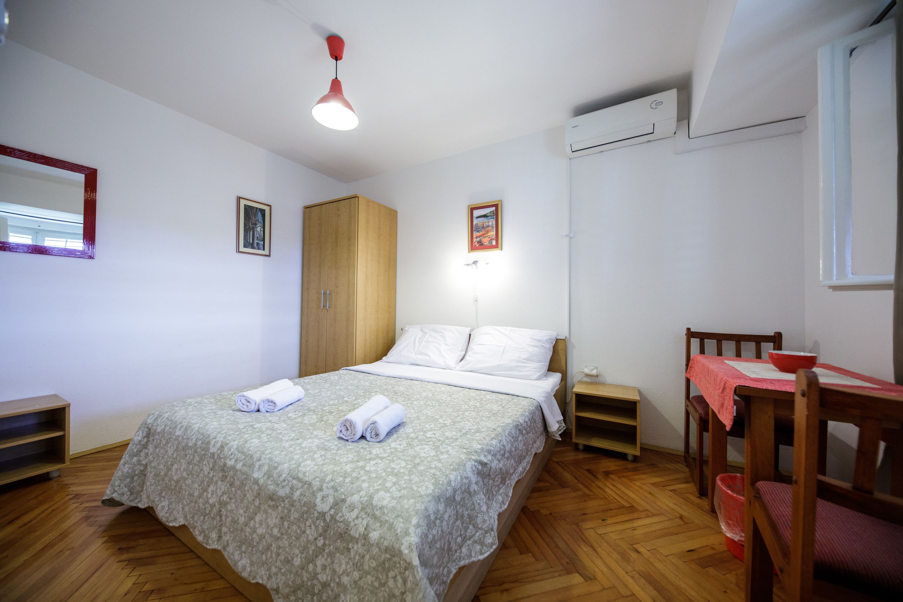 Een slaapkamer met een bed, een houten garderobe, nachtkastje en een kleine eettafel met stoelen.