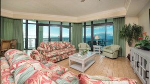 A spacious living room with a large sofa, a coffee table, and two armchairs. The room has large windows with a view of the ocean.
