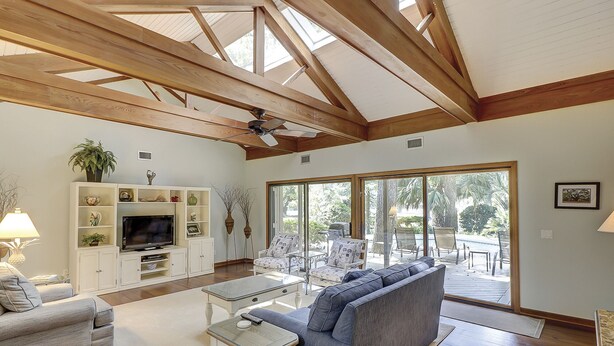 Un salón amplio con techo alto, vigas de madera, televisor, sofá y un gran ventanal corredera que da a un área exterior.