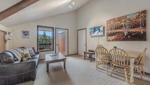 A spacious living room with a sofa, a coffee table, and a dining area with chairs and a table. There are paintings on the walls and a large window with a view of trees. - Ski in Ski out, well appointed 4 bed, 4 bath, with hot tub and lake views! (Sandpoint)