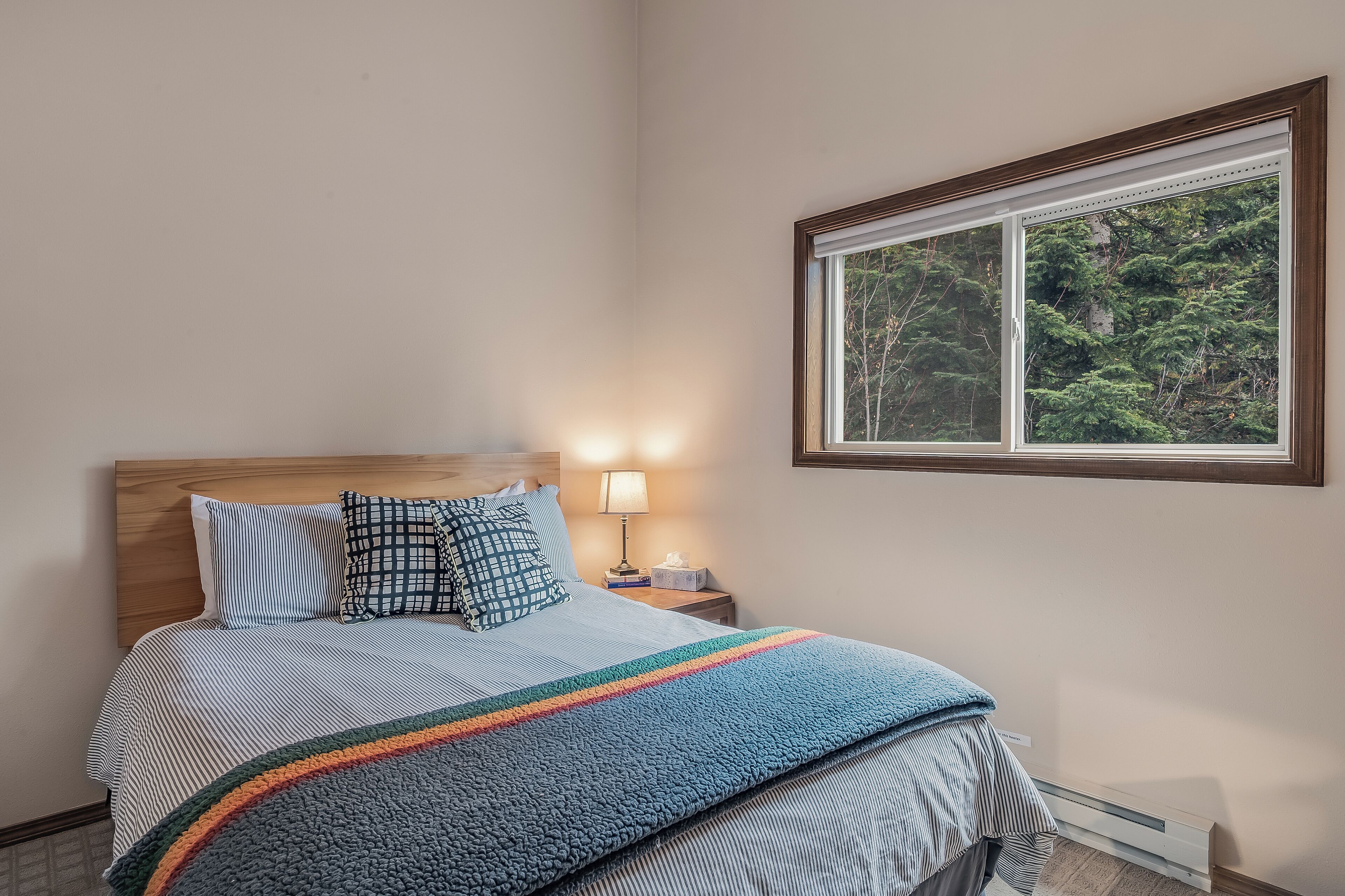 Uma cama bem arrumada com colcha listrada e manta colorida, uma luminária de cabeceira e uma janela com vista para árvores.