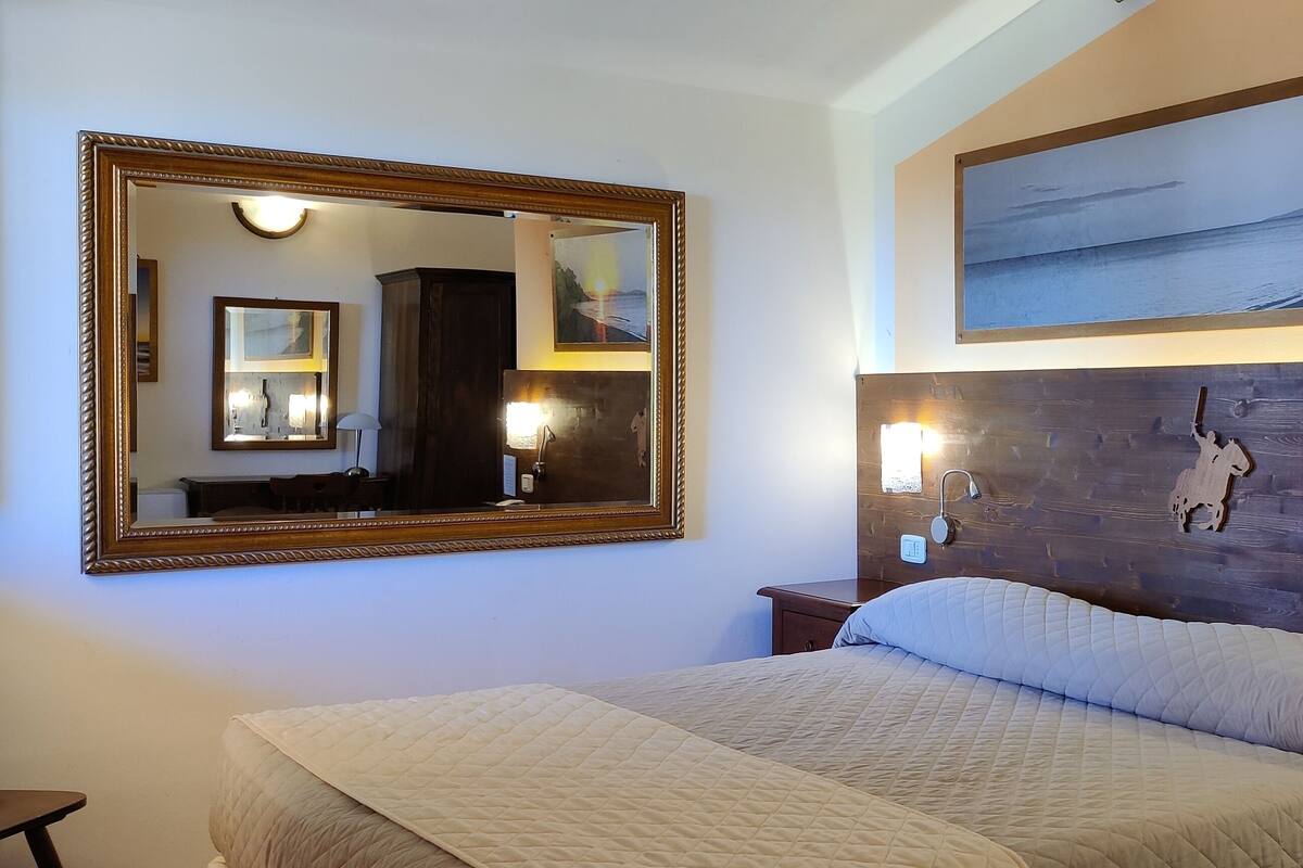 Ein Hotelzimmer mit einem großen Bett, einem hölzernen Kopfteil, einem Nachttisch mit Telefon und Fernbedienung, einem Spiegel und einem Gemälde eines Sonnenuntergangs über dem Meer.