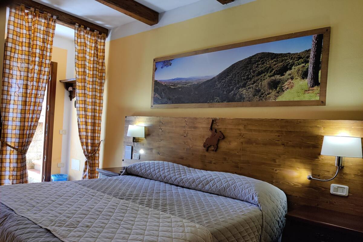 Ein Hotelzimmer mit einem großen Bett, einem Holzkopfende, zwei Nachttischlampen, einem aufgehängten Landschaftsbild und karierten Vorhängen.