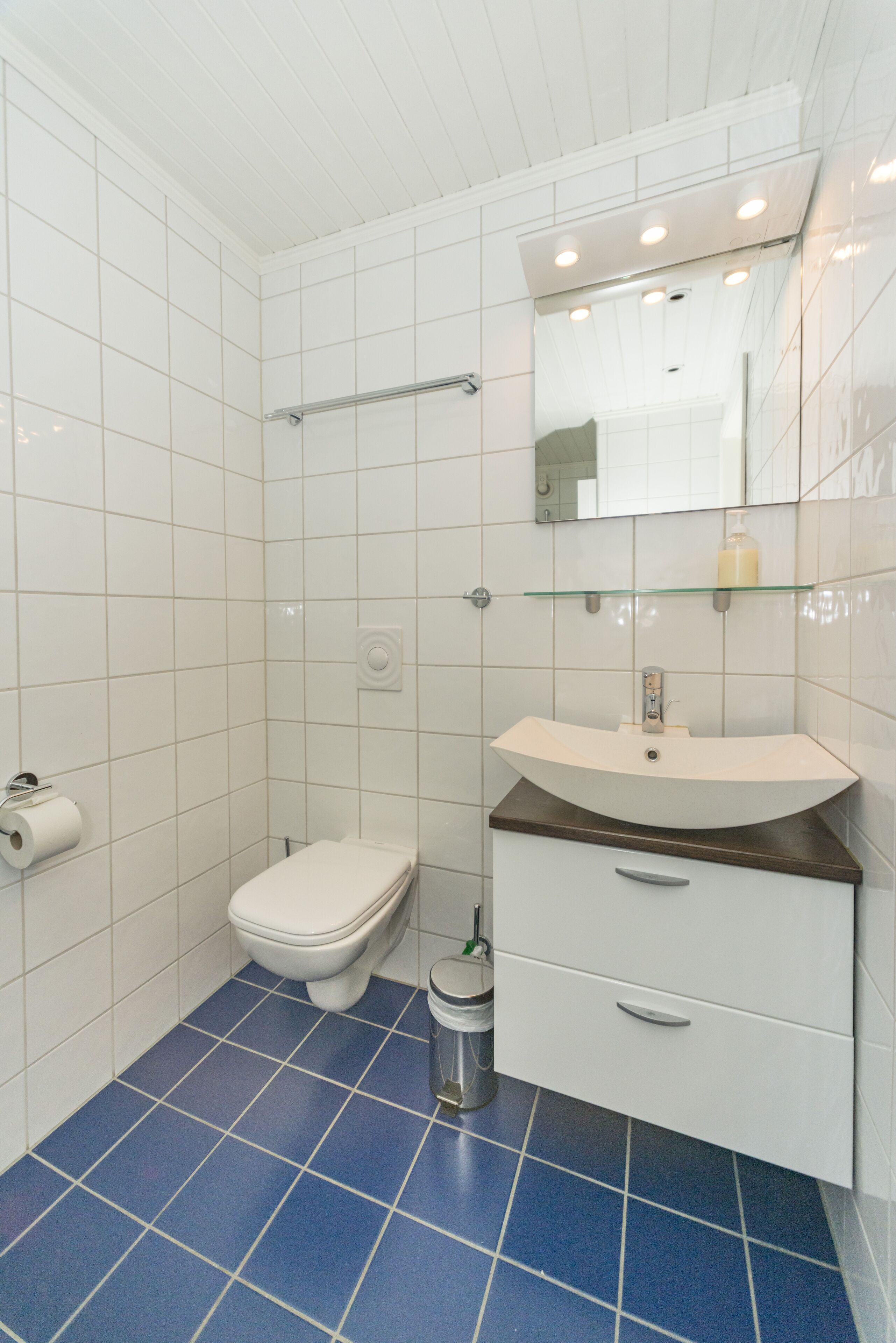 A bathroom with white tiles, a blue-tiled floor, a white toilet, a glass shelf with a bottle, and a white sink with a brown countertop.