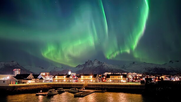 A coastal town with a dock, buildings, and snow-capped mountains under a night sky with the Northern Lights.