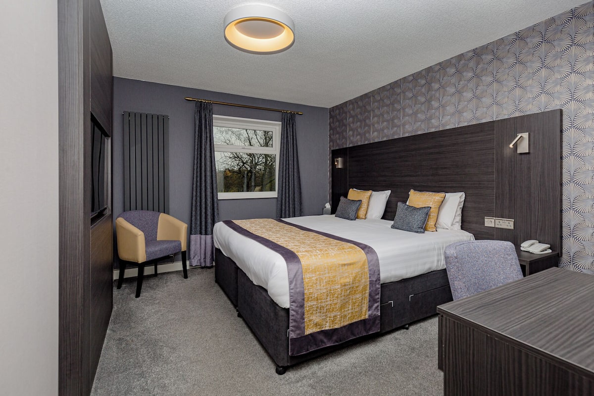 Una habitación de hotel moderna con una cama grande, un escritorio, una silla y un ventanal con cortinas.