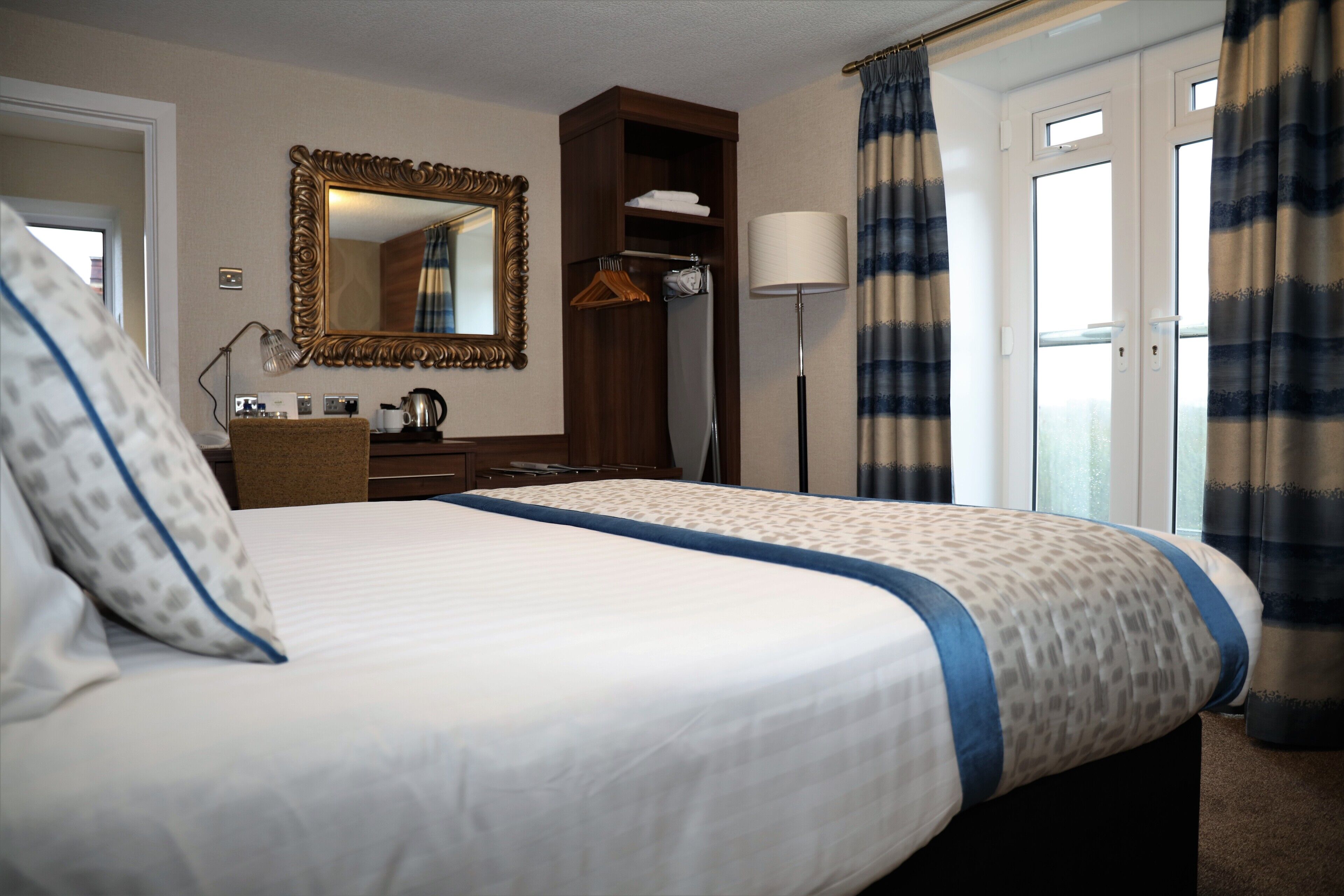 A hotel room with a large bed, a wardrobe, a mirror, a bedside table with a lamp, and a view of the outside through a door.