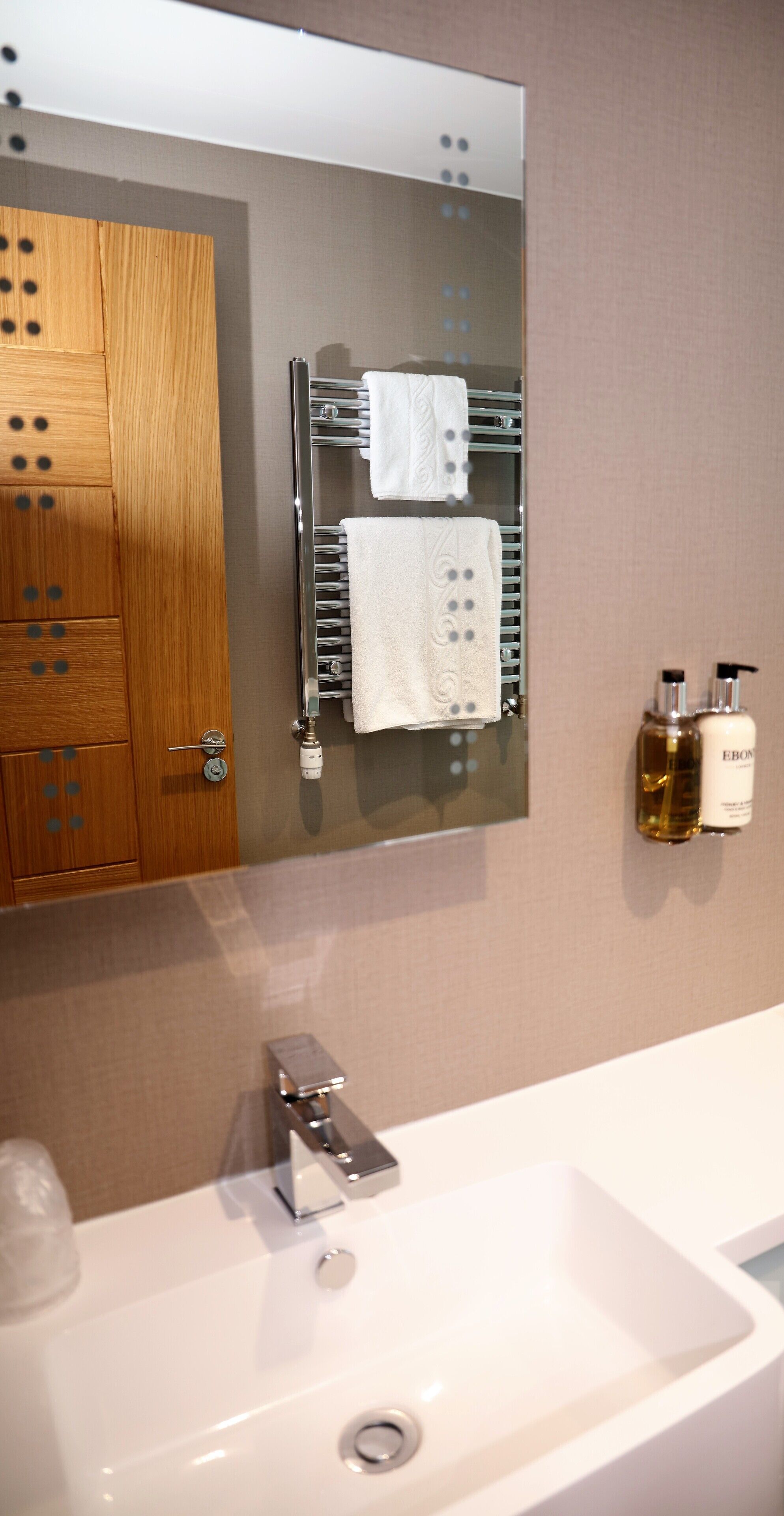 A bathroom with a white sink, a mirror, a towel rack with two white towels, and two bottles of Ebon body wash on the wall.