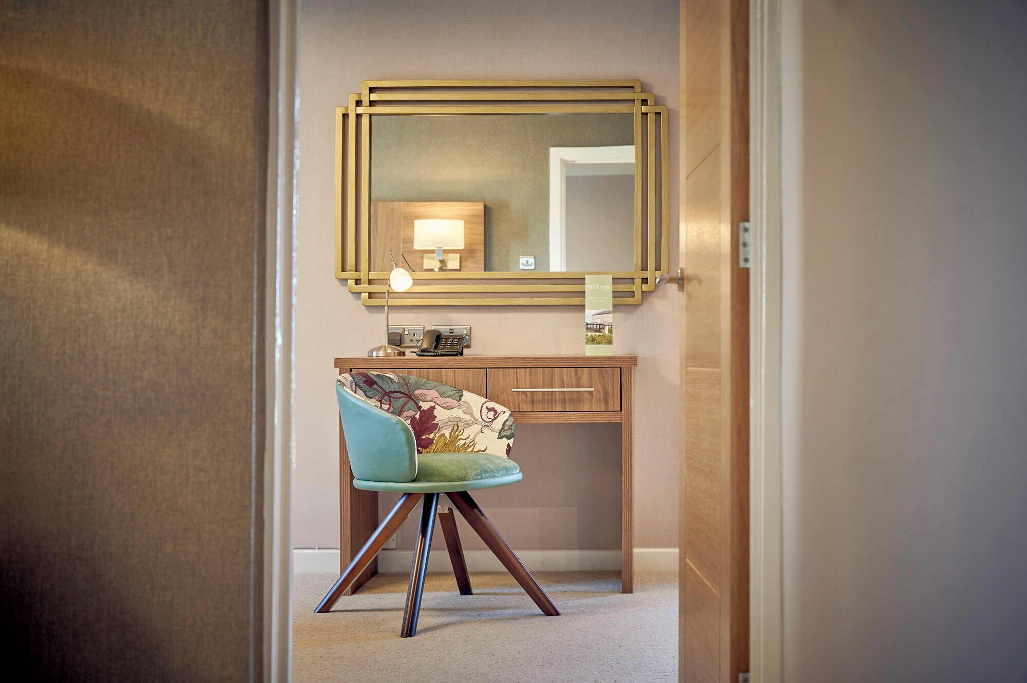 Ein Hotelzimmer mit einem Holz-Waschtisch, einem Spiegel, einem Schreibtisch mit Lampe und einem stuhl mit Blumenmuster.