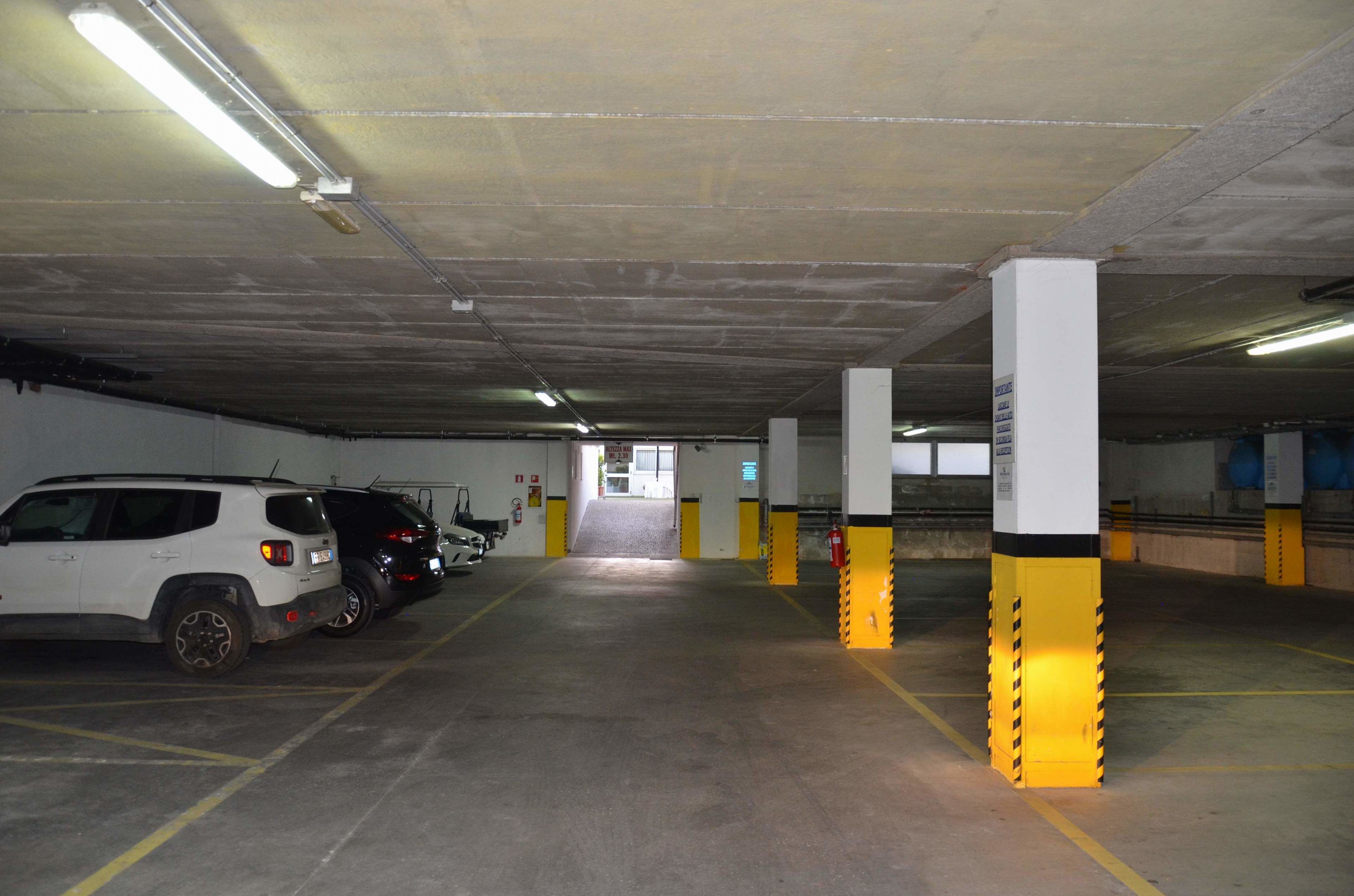 Un parcheggio sotterraneo con pilastri in cemento, luci fluorescenti e alcune auto parcheggiate.