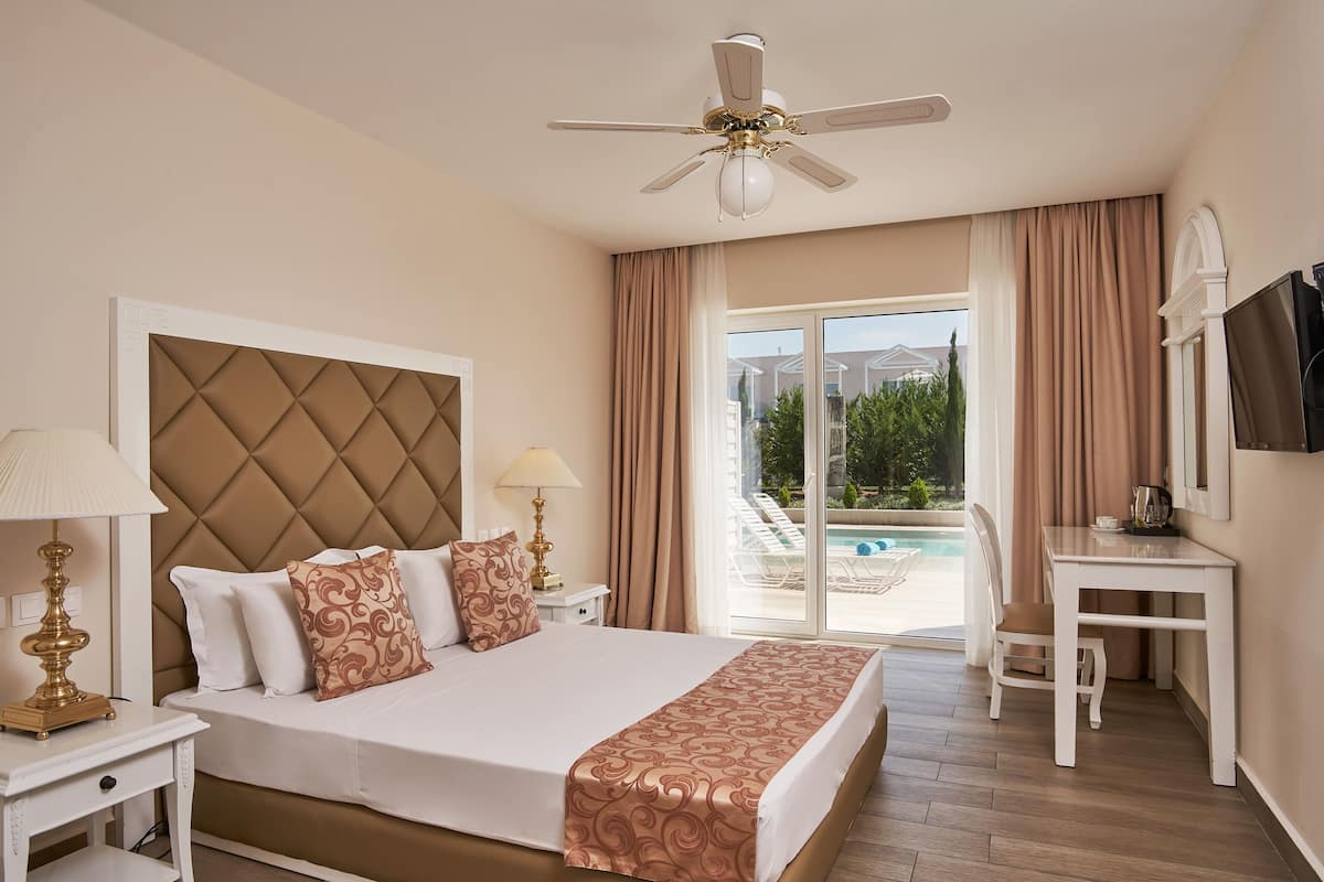 Habitación de hotel con cama, escritorio, silla, ventilador de techo y vista a una piscina.