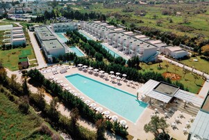 Μια αεροφωτογραφία ενός θέρετρου με μια μεγάλη πισίνα, η οποία περιβάλλεται από ξαπλώστρες και ομπρέλες, καθώς και από πολλά κτίρια του ξενοδοχείου και διαμορφωμένους κήπους.