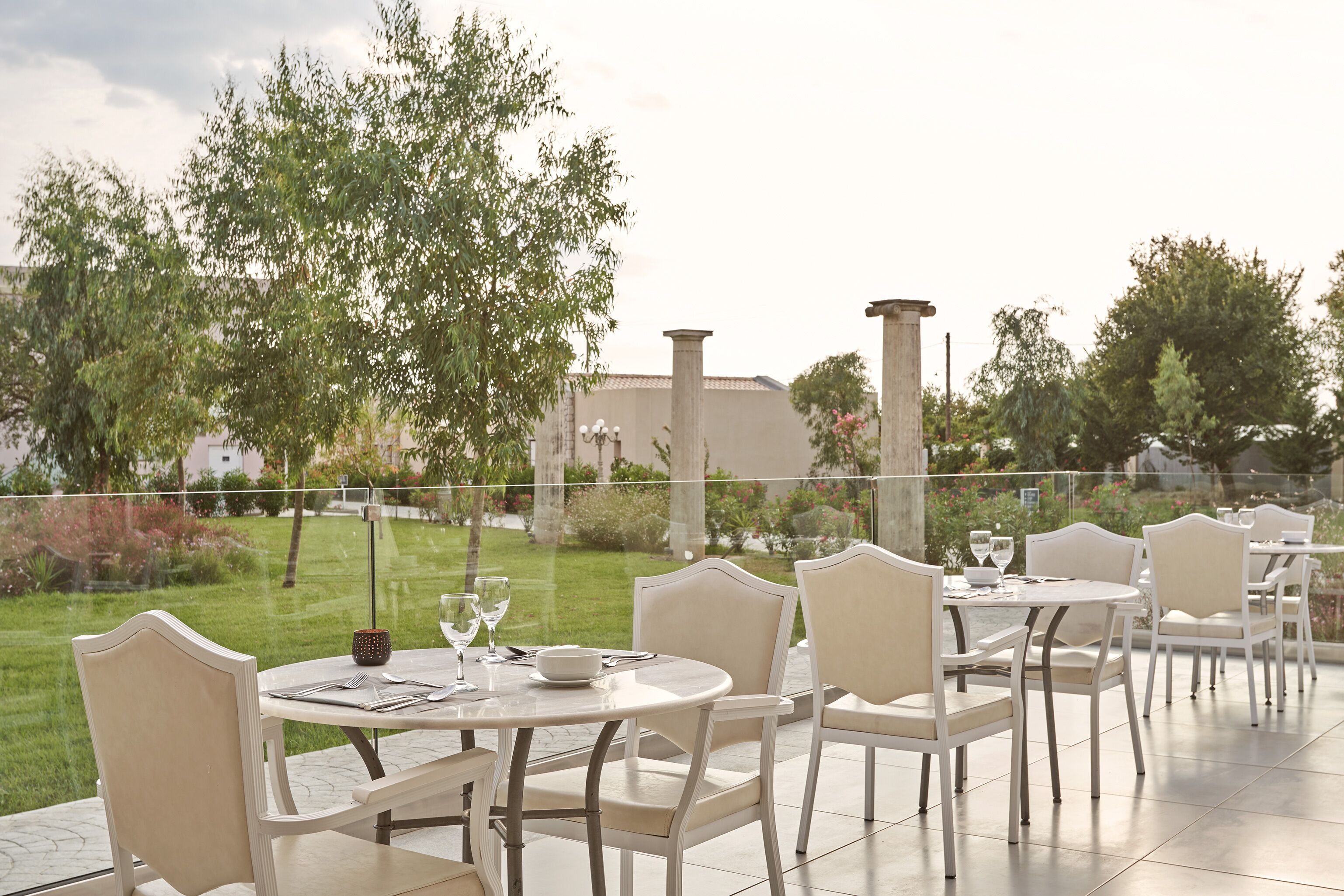 A patio with round tables and white chairs, overlooking a garden with trees and a building in the background.