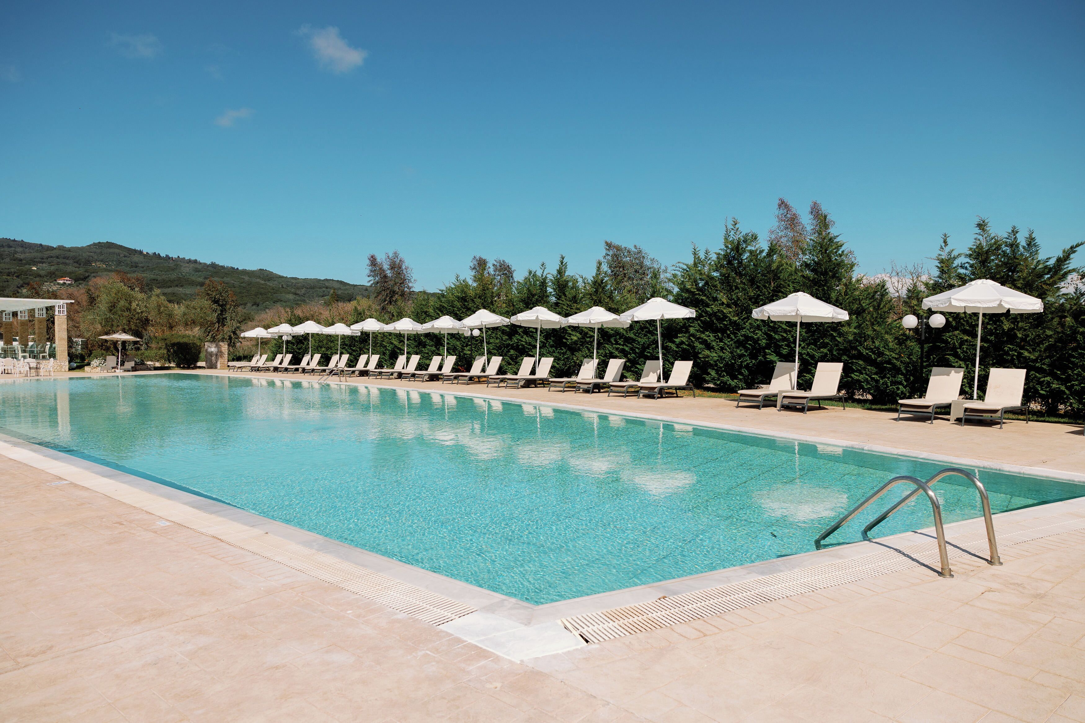 Piscina com espreguiçadeiras e guarda-sóis, cercada por árvores e colinas.