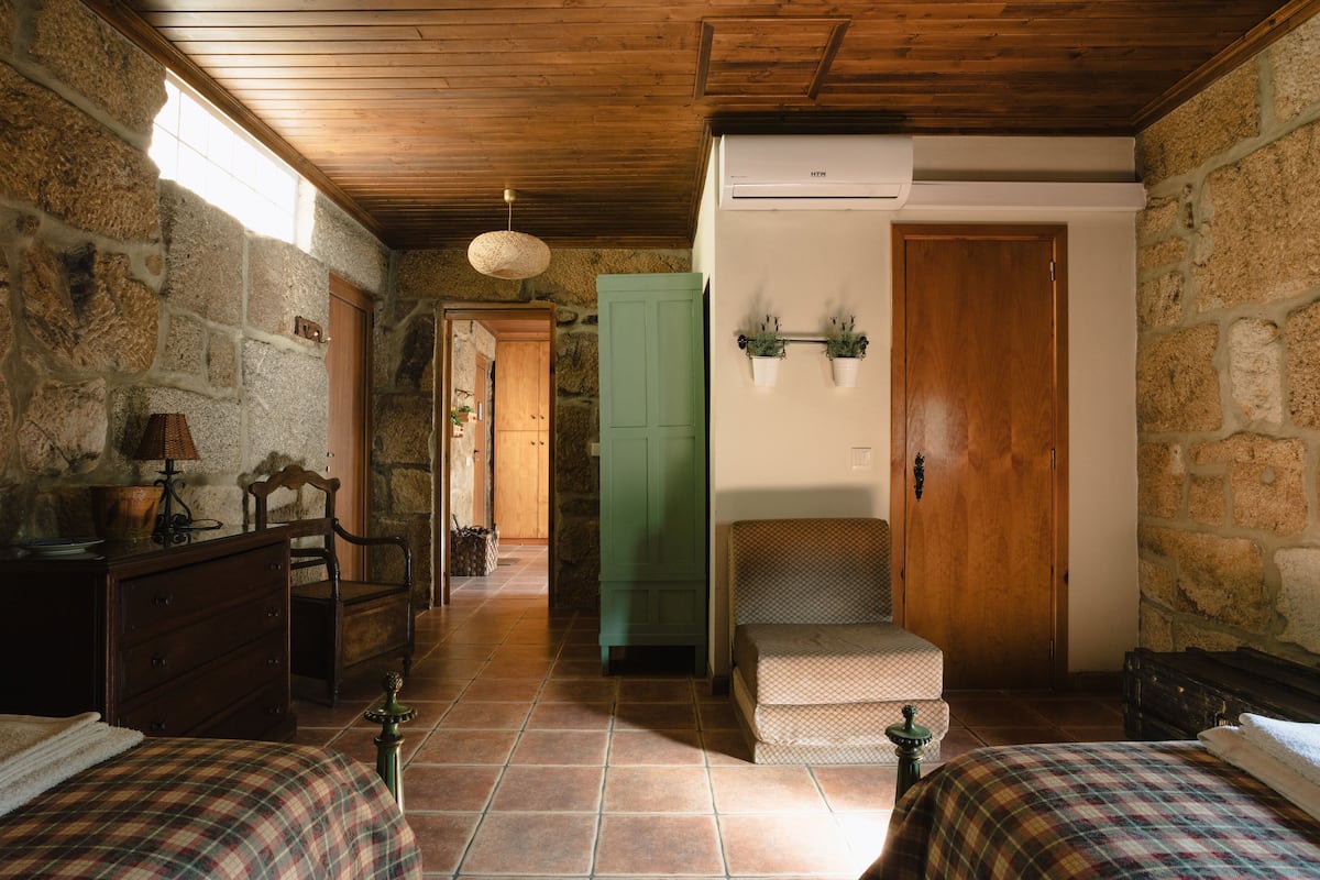 A rustic room with stone walls, wooden ceiling, two beds with plaid bedding, a wooden dresser, a lamp, a green cabinet, and a staircase.