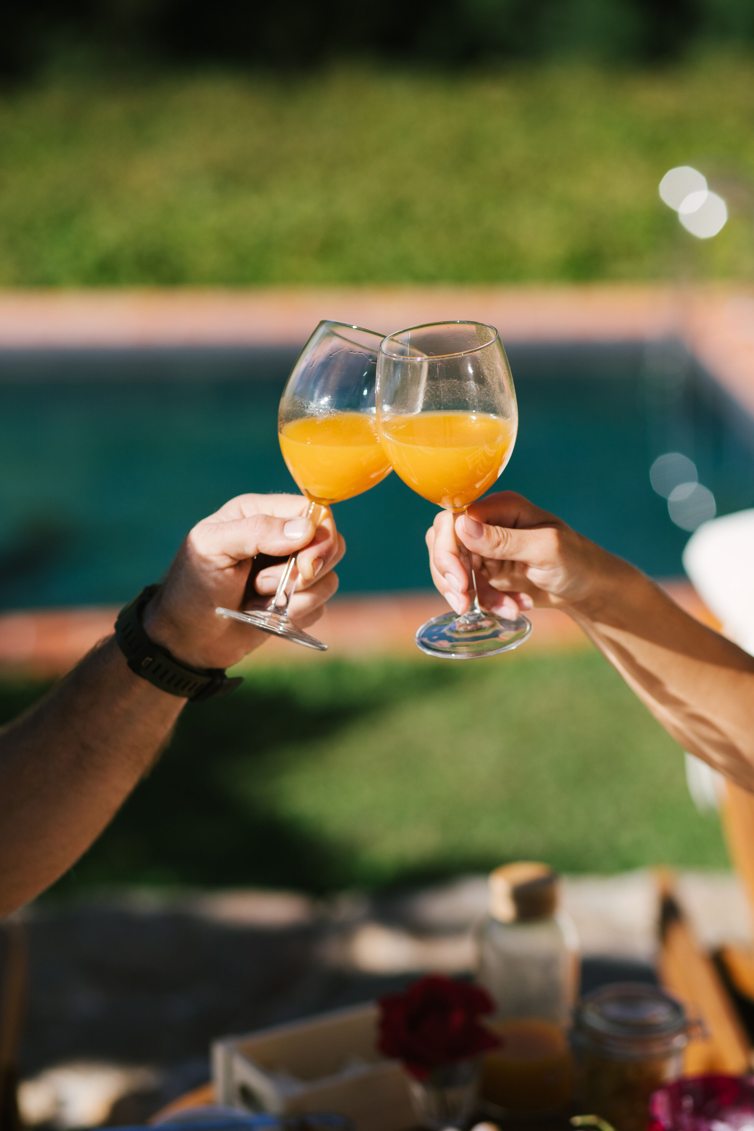 Two people toasting with orange drinks at a table with a pool in the background.