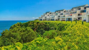 Un hôtel en bord de mer doté de nombreux balcons donnant sur l’océan, d’une végétation luxuriante à l’avant et d’un ciel bleu dégagé.