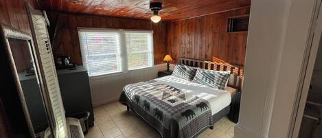 Un dormitorio con techo de madera, una cama con colcha, una mesita de noche, una lámpara y una ventana con persianas.