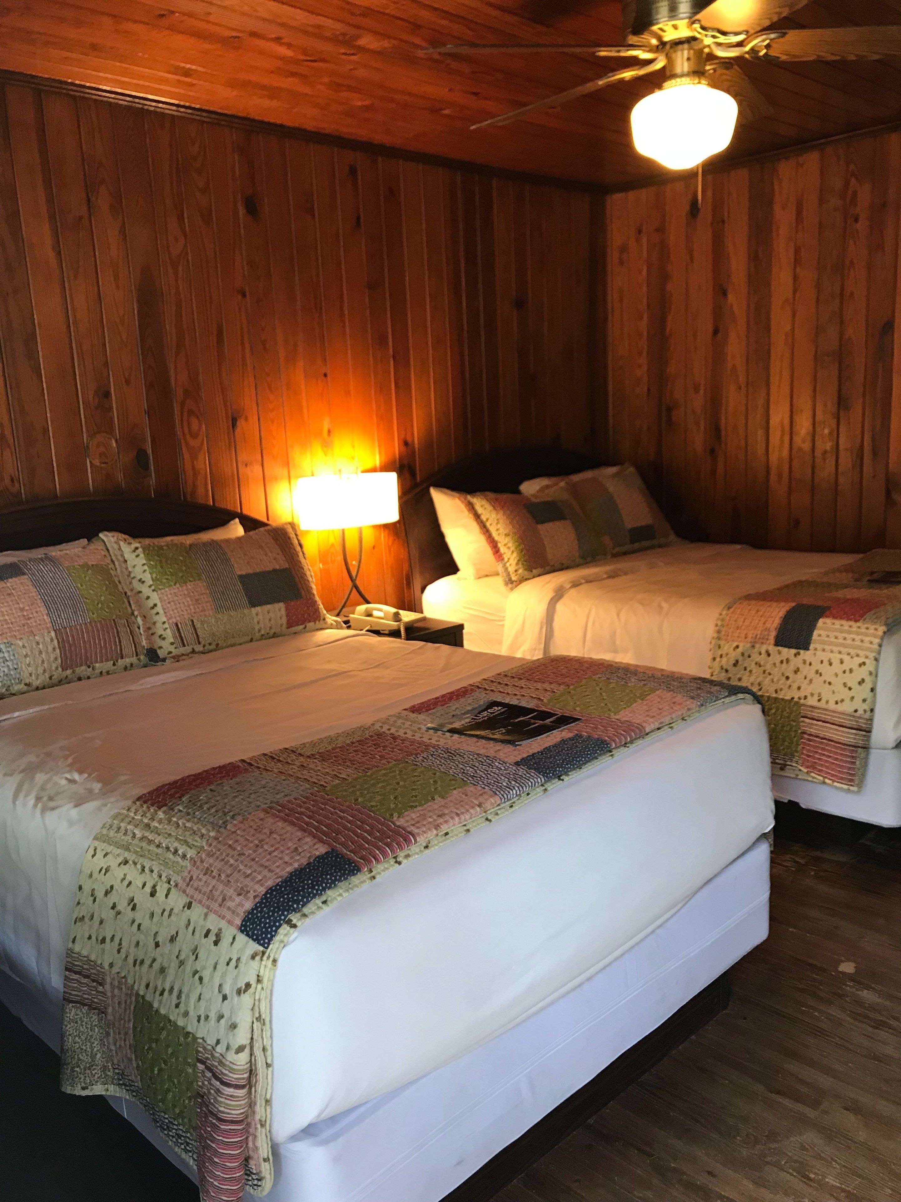 A room with two beds, wooden walls, a ceiling fan, and a wall-mounted lamp.