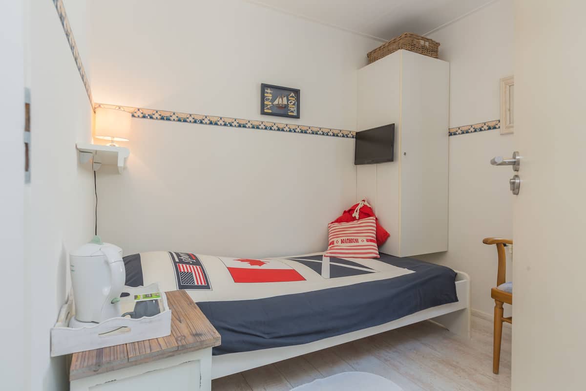 A small, neatly arranged room with a bed, a desk with a kettle, a chair, and a TV mounted on the wall.