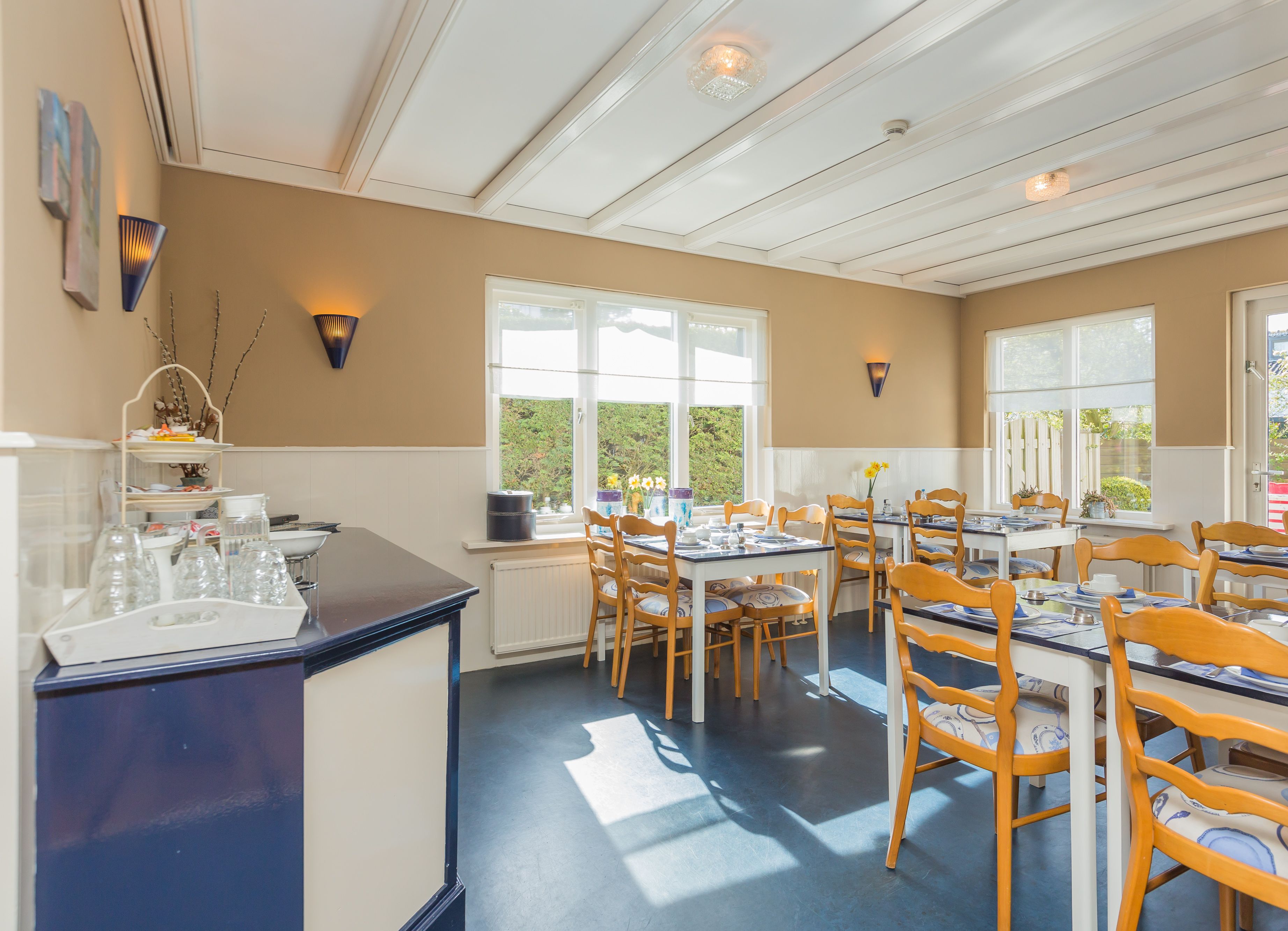 A dining area with multiple tables and chairs, a kitchen counter with glassware, and large windows providing natural light.