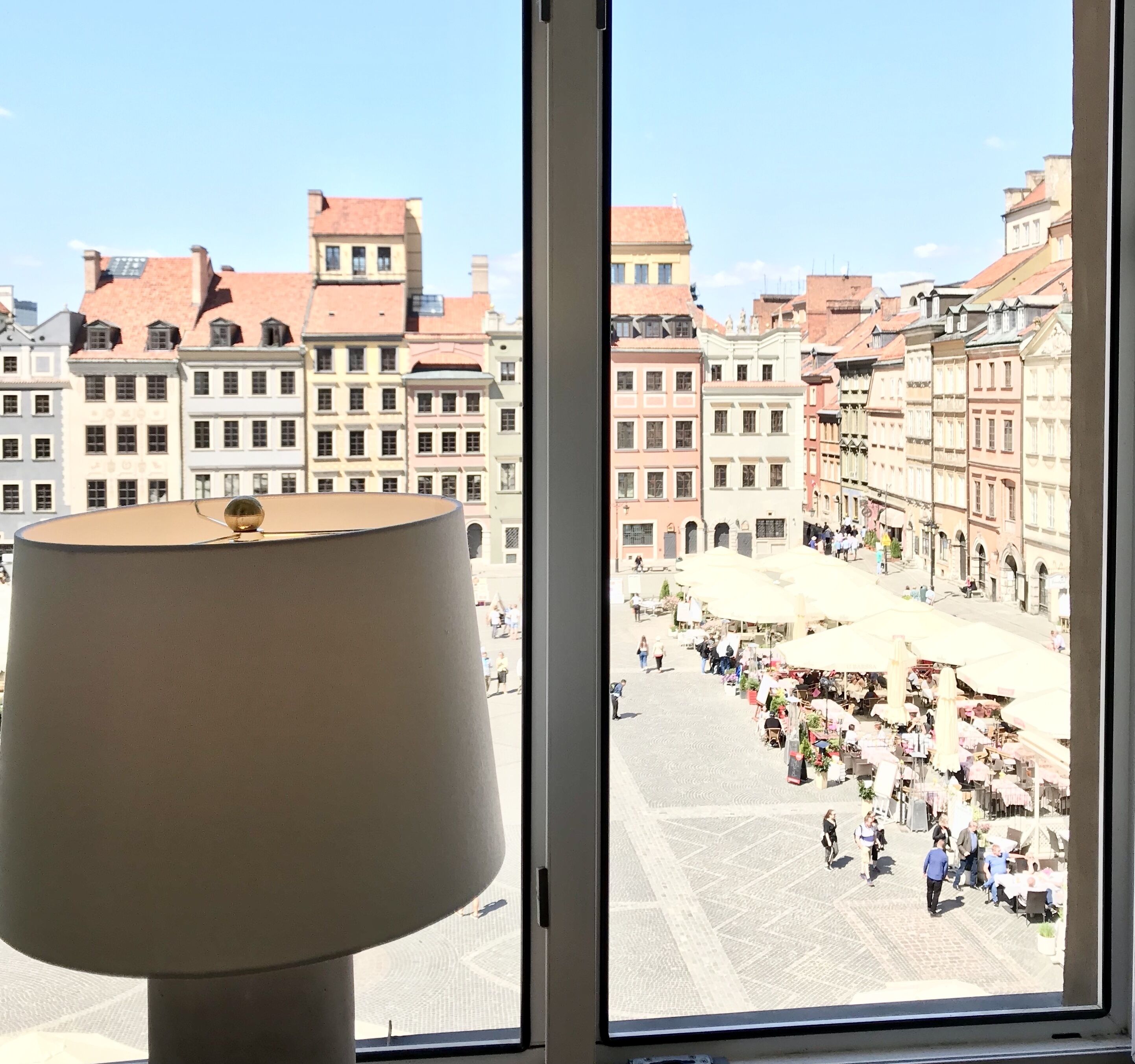 Una ventana con vista a una plaza urbana, edificios y gente.