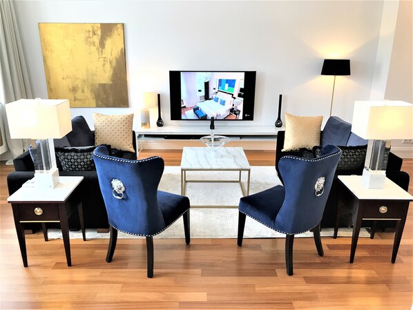 Een moderne woonkamer met een flatscreen televisie, twee blauwe fauteuils, een salontafel en twee bijzettafels met lampen.