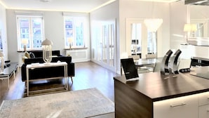Un salon moderne avec un canapé noir, une table basse blanche et un tapis. On y trouve également un coin repas avec une table et des chaises, ainsi qu’une cuisine avec un comptoir et un luminaire suspendu.