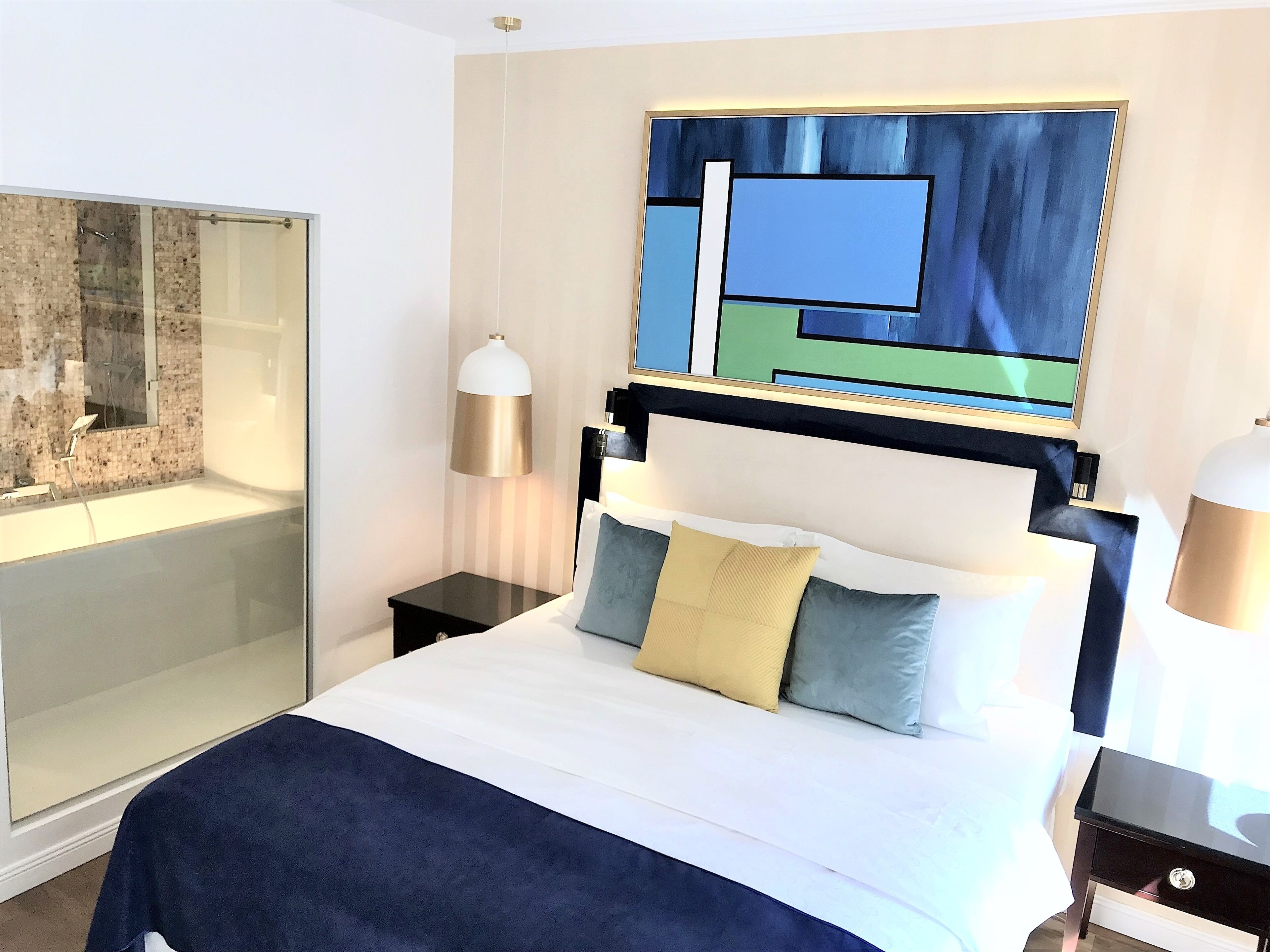 Habitación de hotel con cama, mesitas de noche, cabina de ducha de vidrio y un cuadro abstracto grande en la pared.