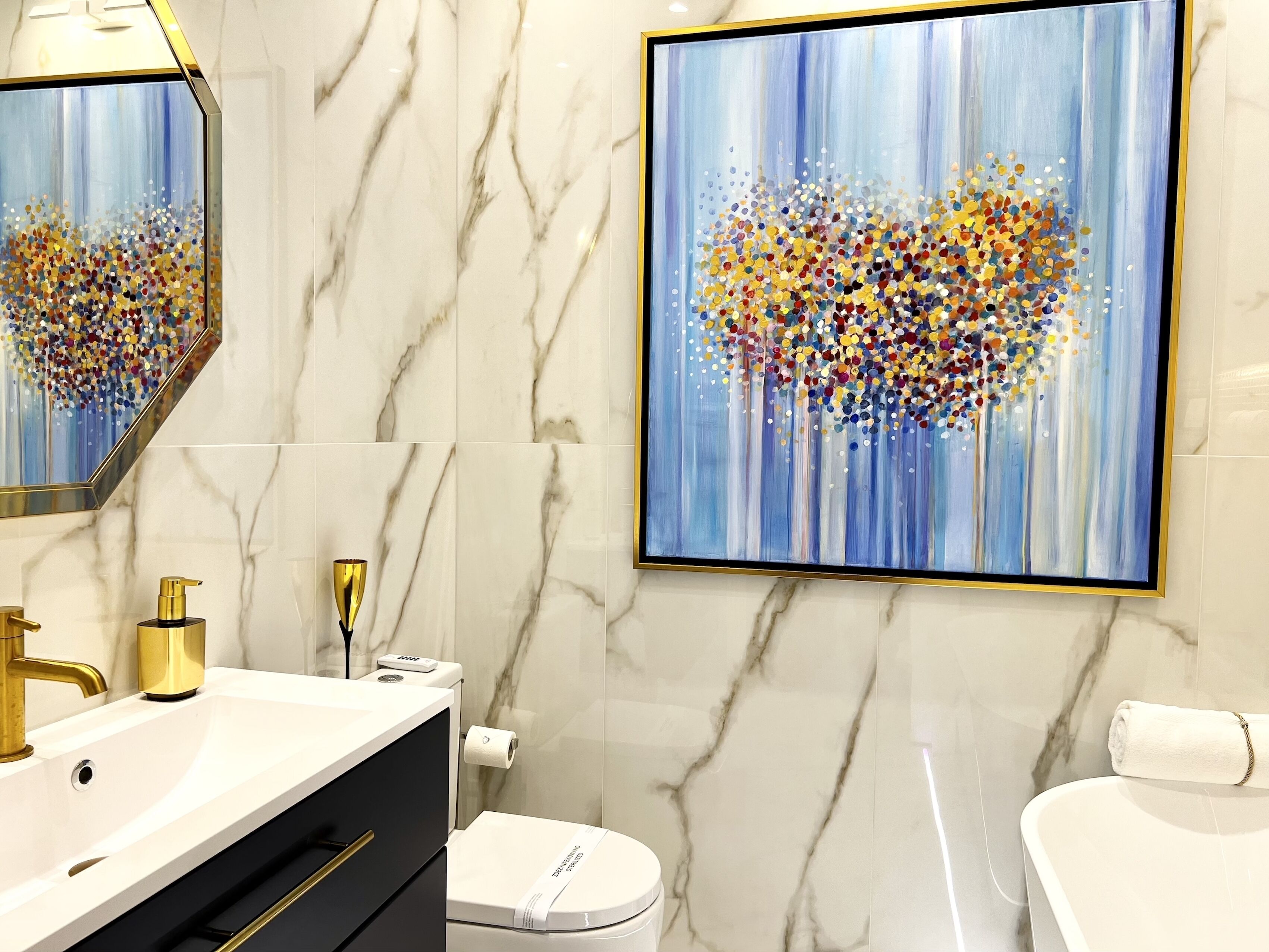 A modern bathroom with a marble backsplash, a white sink with gold fixtures, a toilet, and a large abstract painting on the wall.