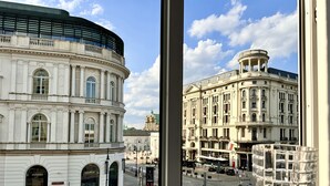 Vue d’un bâtiment historique à l’architecture classique, avec ses colonnes et ses fenêtres en arc de cercle, prise depuis une fenêtre donnant sur une rue animée par des piétons et bordée de voitures garées.