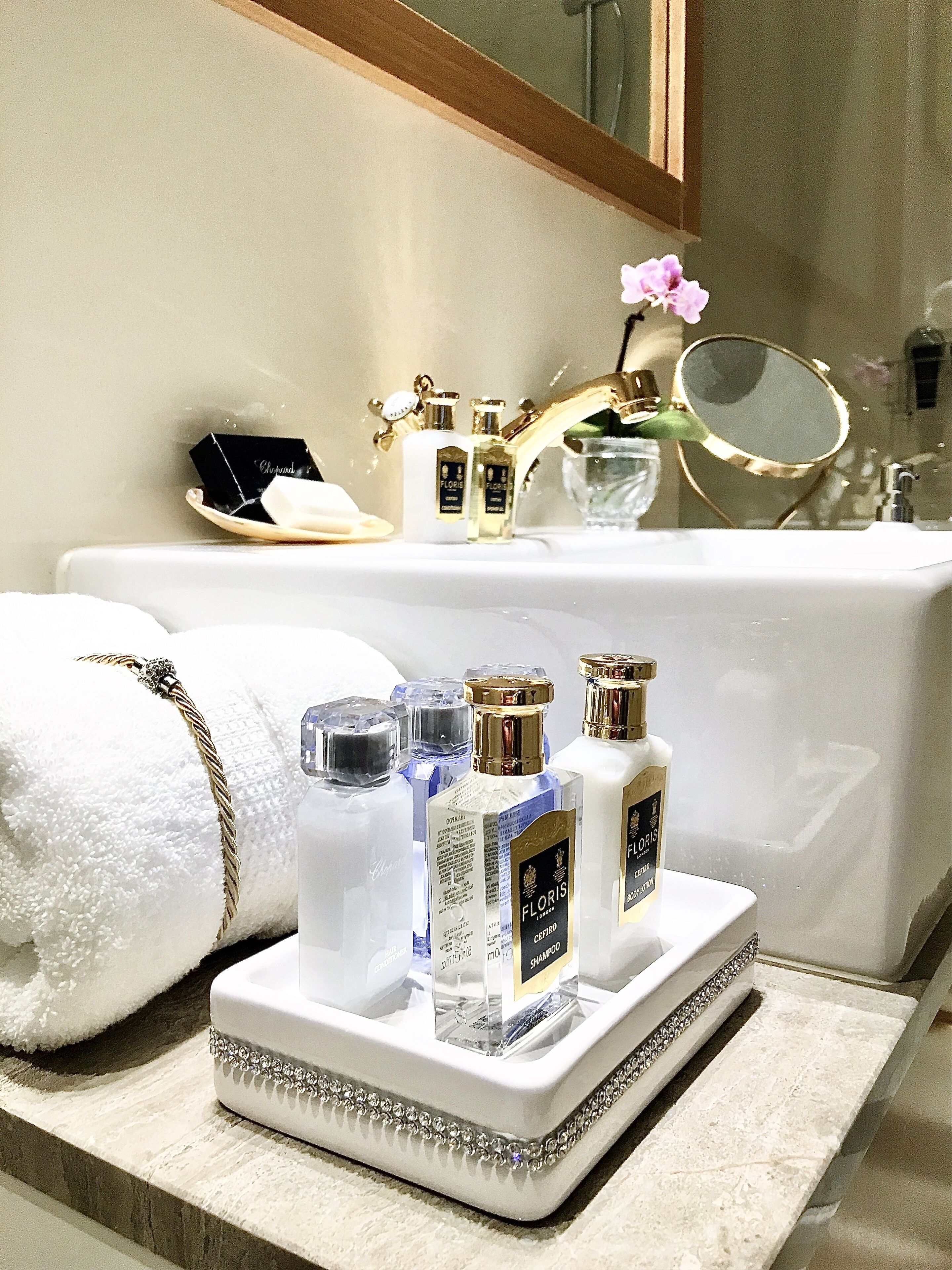 Een wastafelmeubel met een witte wasbak, een gouden kraan en een spiegel. Op het aanrecht staan verschillende flesjes parfum en een witte handdoek.