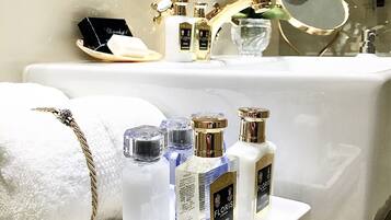 Een wastafelmeubel met een witte wasbak, een gouden kraan en een spiegel. Op het aanrecht staan verschillende flesjes parfum en een witte handdoek.